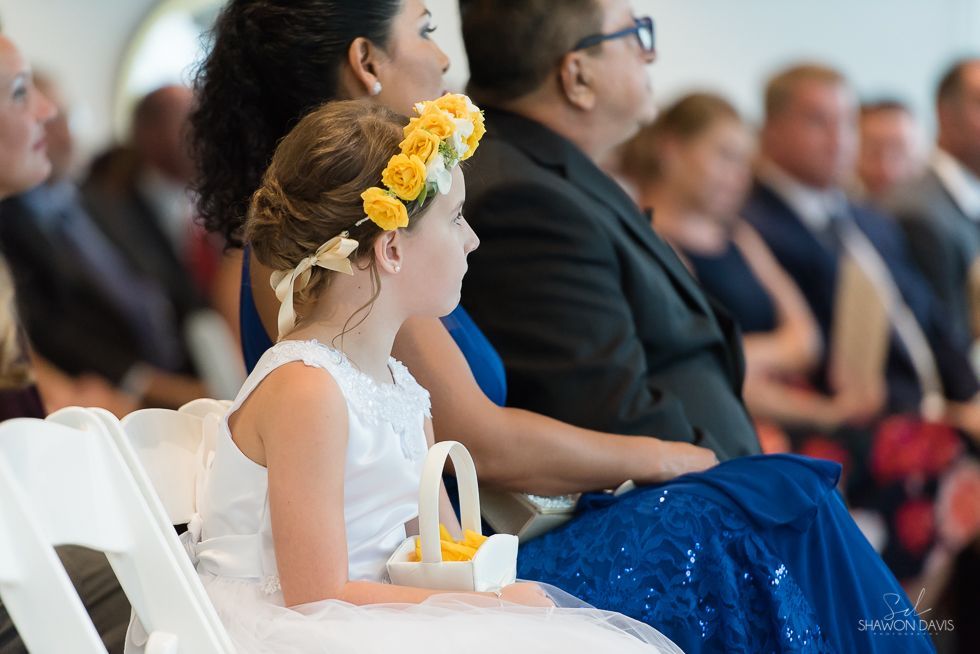 Girl in white dress with yellow flower crown seated at an indoor ceremony, holding a small candle