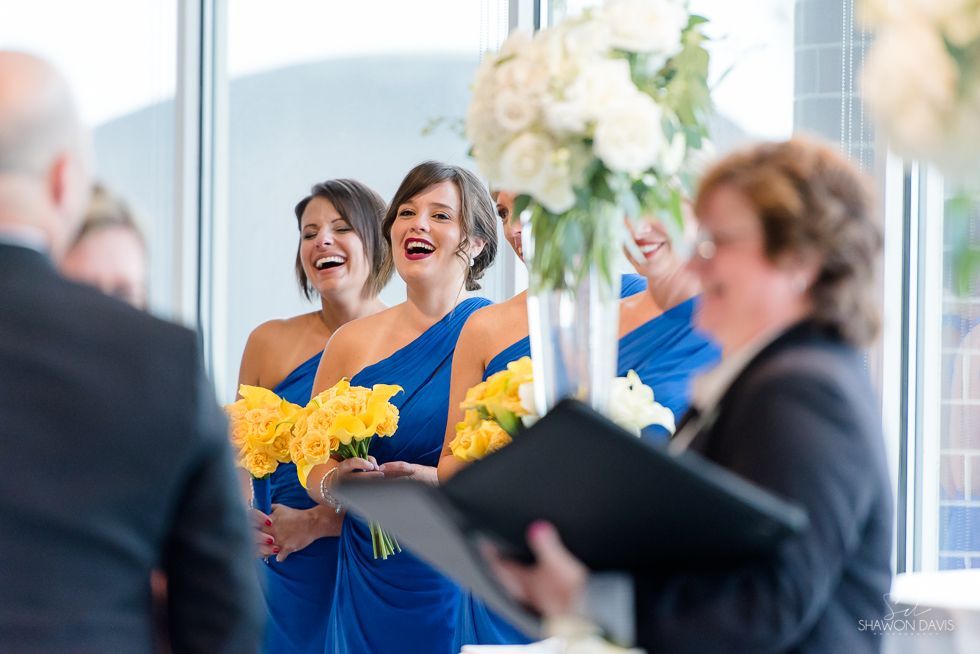 Bridesmaids in blue dresses holding yellow bouquets smile at a wedding ceremony in a bright venue