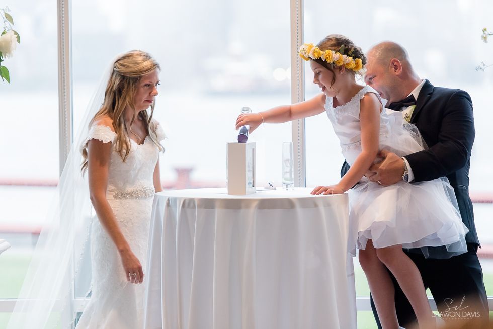 Bride and groom lighting a unity candle beside a woman in a white dress at a bright wedding table