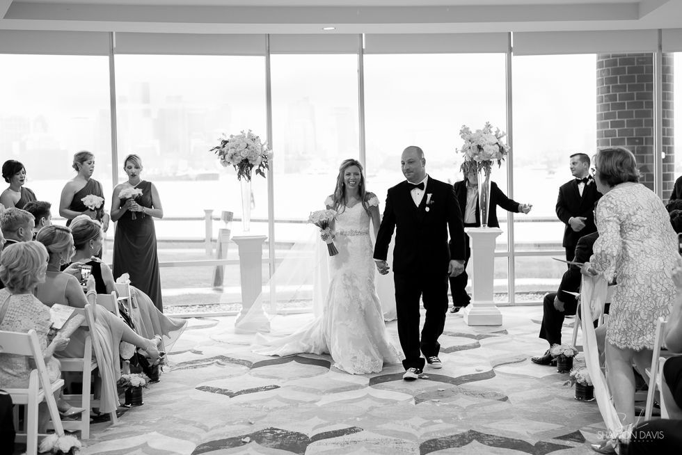 Bride and groom walk down the aisle in a bright wedding venue, guests seated on both sides.