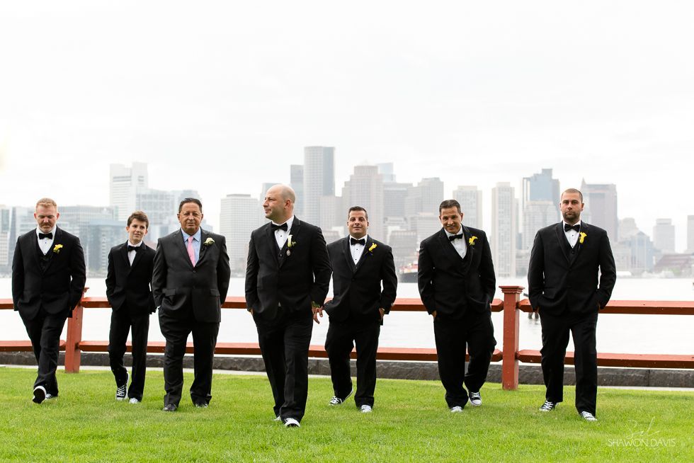 Six men in black suits walk across a green lawn with a city skyline in the background.