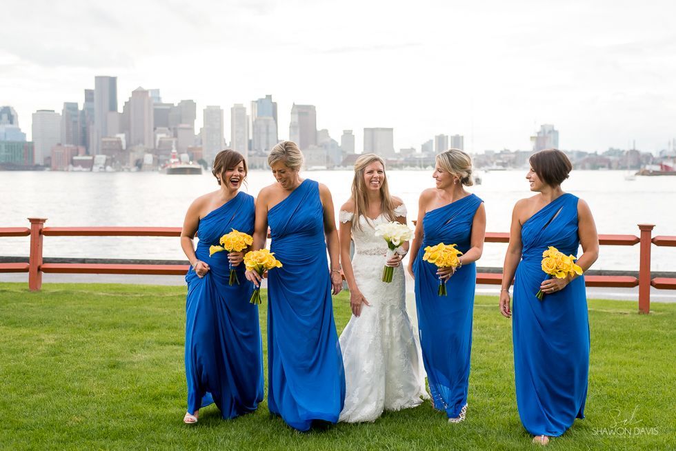 Five women in blue and white dresses stand by a waterfront railing with a city skyline behind them.