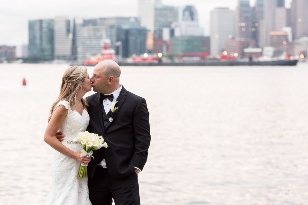 Bride and groom embracing by the water, with a city skyline in the background.