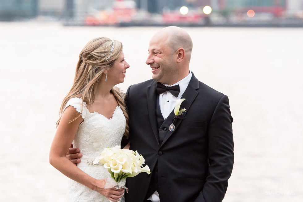 Bride and groom smiling on a beach, holding hands and bouquet in wedding attire