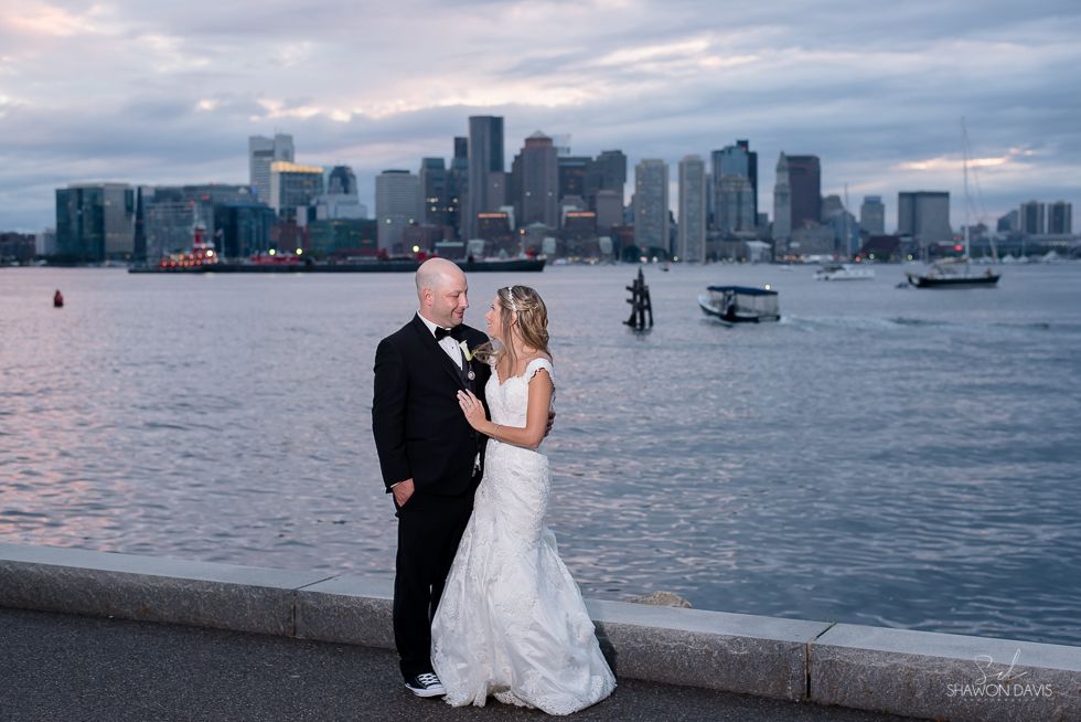 Bride and groom in wedding attire by the water with a city skyline at dusk