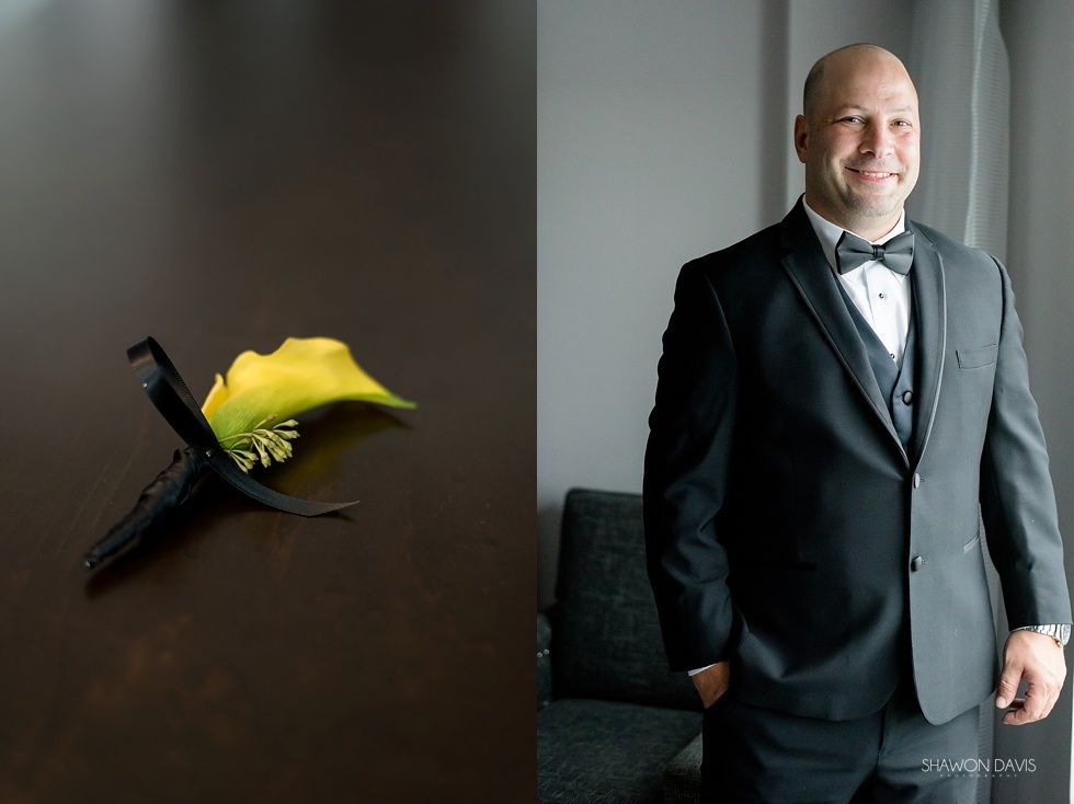Man in a black tuxedo stands smiling beside a wall sculpture with a yellow flower against a gray background