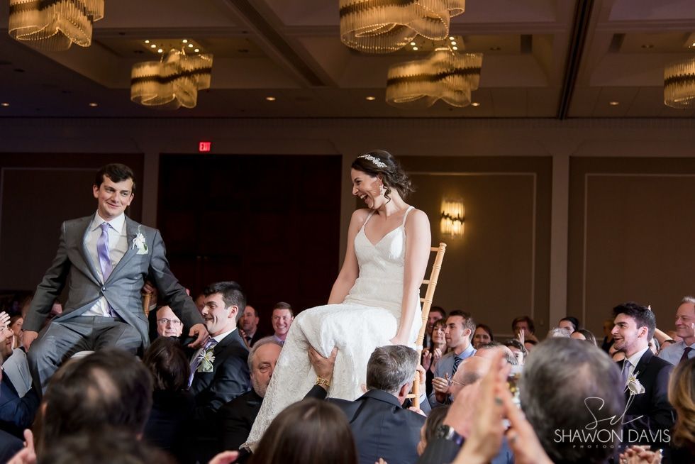 Bride and groom smiling at their wedding reception, walking through a crowd of seated guests.