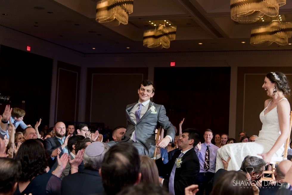 Groom in gray suit walks down an aisle as guests applaud, while bride stands smiling nearby at a reception