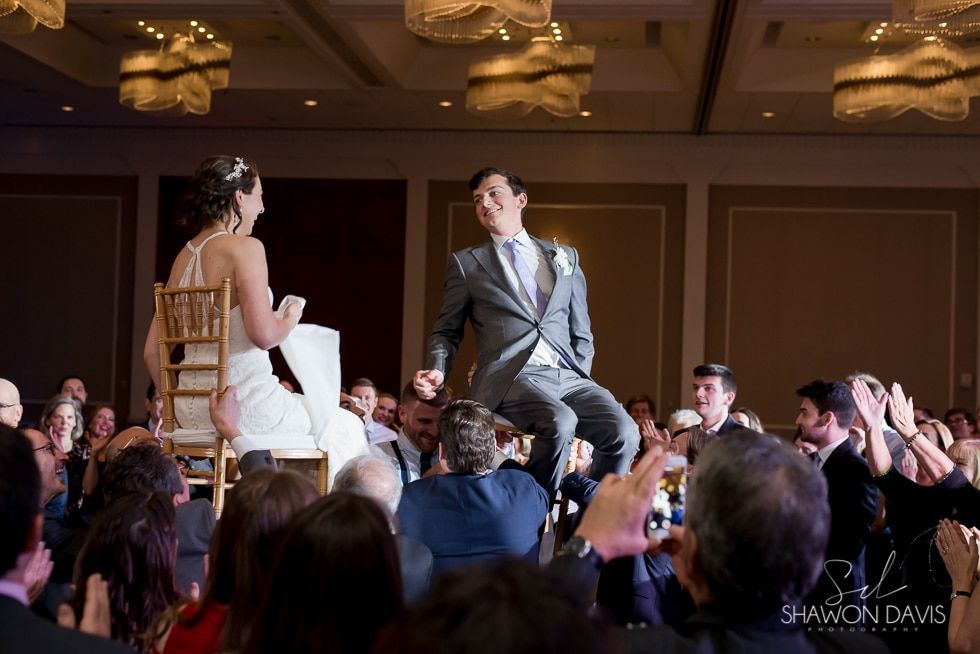 Bride and groom seated on chairs during a wedding reception, smiling at guests raising hands around them
