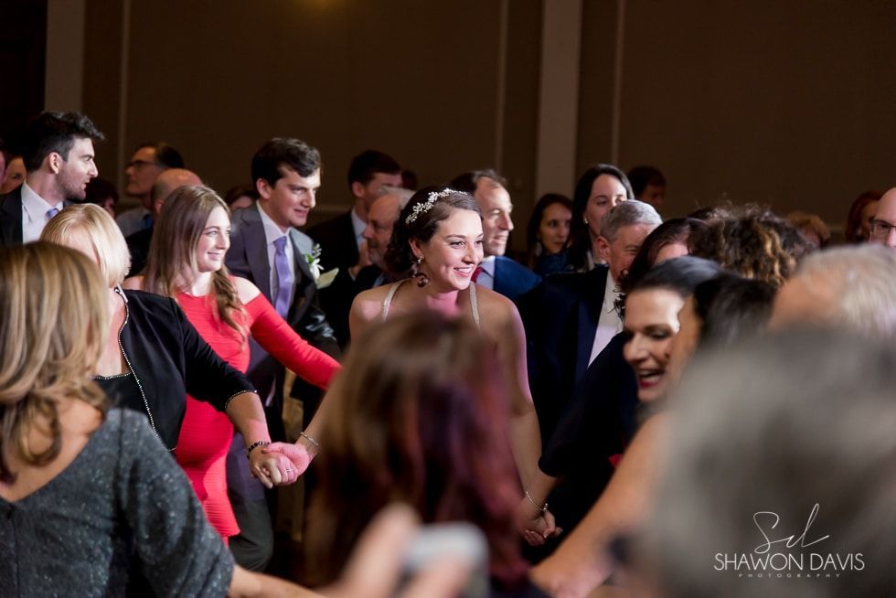 Guests dancing at a lively indoor event, with a smiling woman in a pink dress at center.
