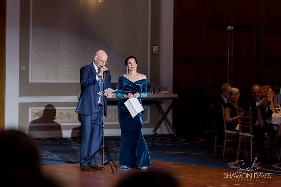 Two presenters on a dimly lit stage, one speaking at a microphone, the other holding notes.