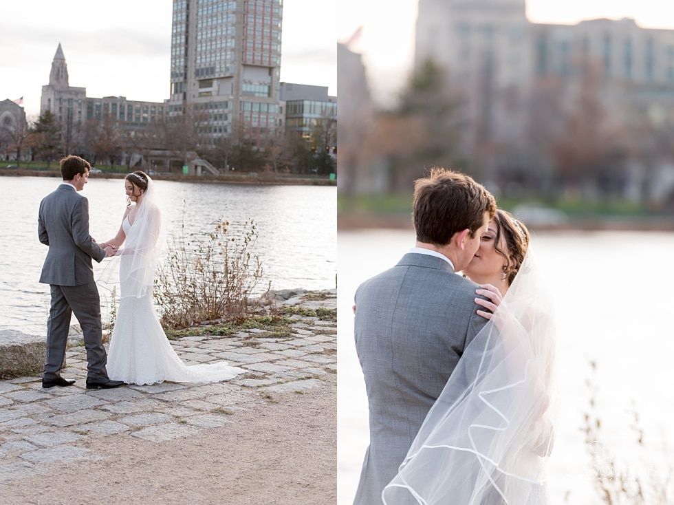 Couple in wedding attire by a river, embracing with city buildings in the background.