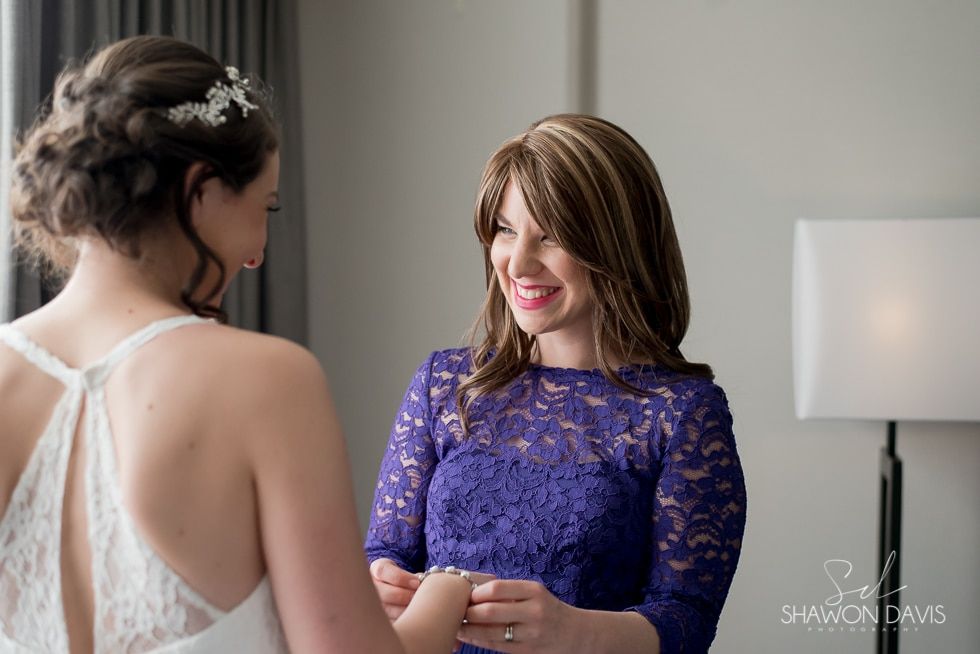 Two women smiling and talking, one in a purple lace dress and one in a white gown, in a bright room