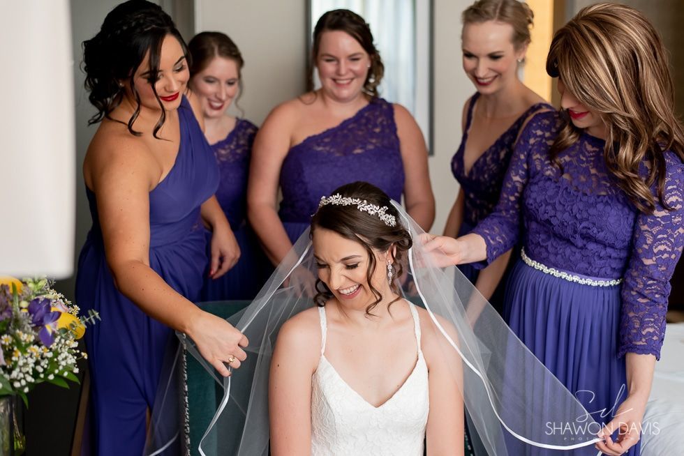 Bride seated while five women in purple dresses adjust her veil in a bridal room
