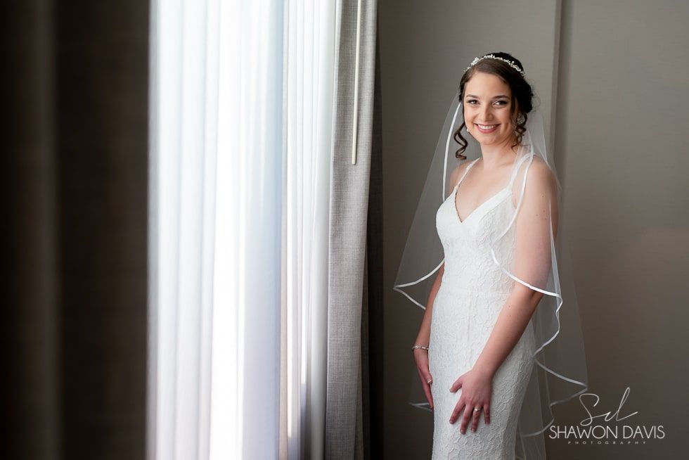 Bride in a white lace gown standing by a sunlit window, smiling softly