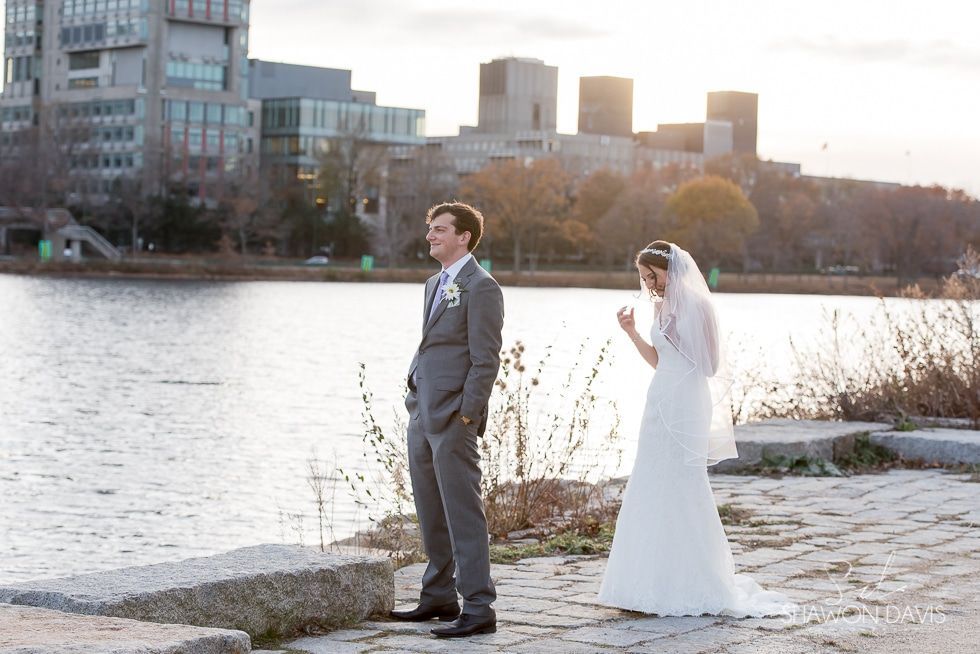 Bride and groom standing by a riverside at sunset, with city buildings in the background.