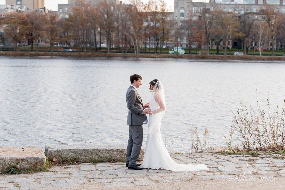 Bride and groom holding hands by a riverbank, with city trees in the background