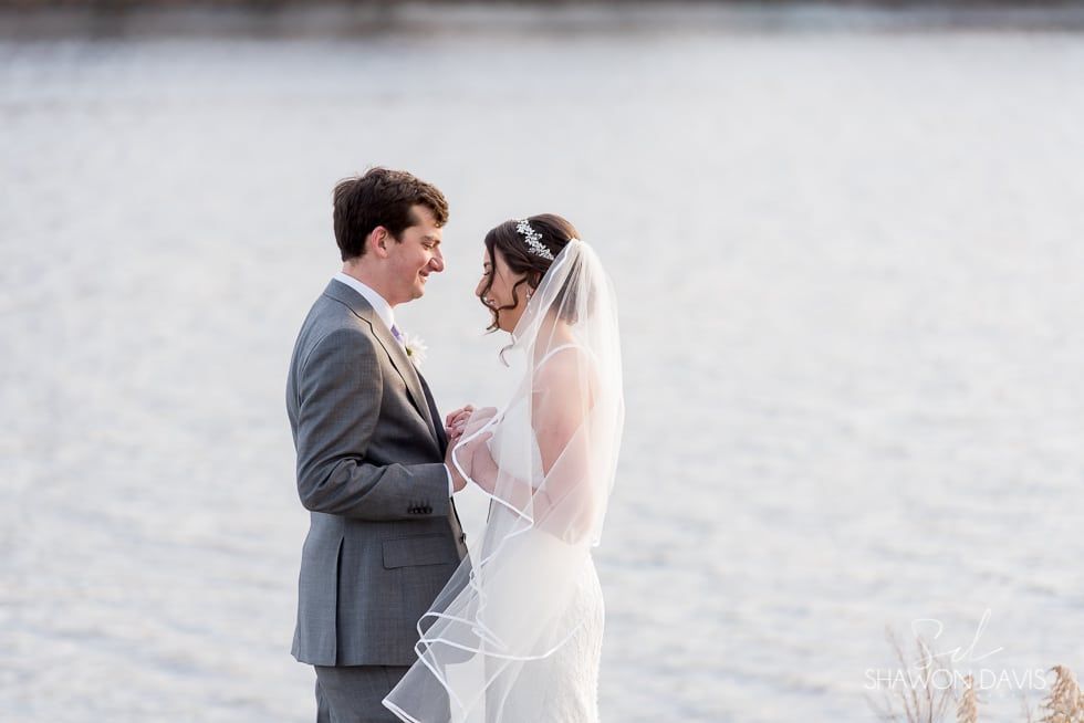 Bride and groom facing each other by a calm lake, holding hands in wedding attire