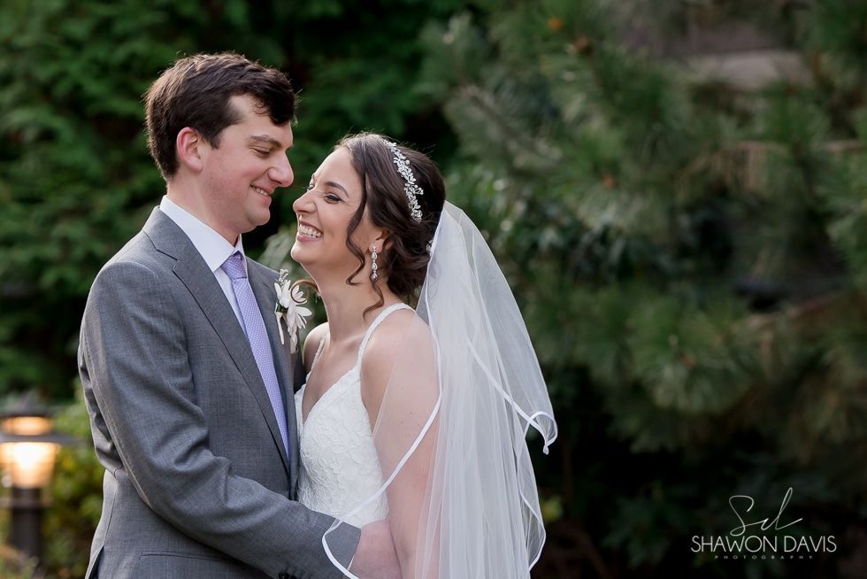 Bride and groom smiling in a garden, holding each other in wedding attire and veil