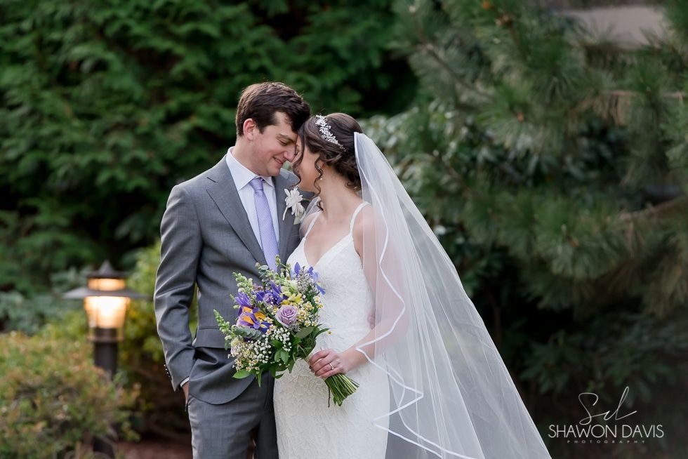Bride and groom embracing in a garden, with bouquet and veil in soft greenery.