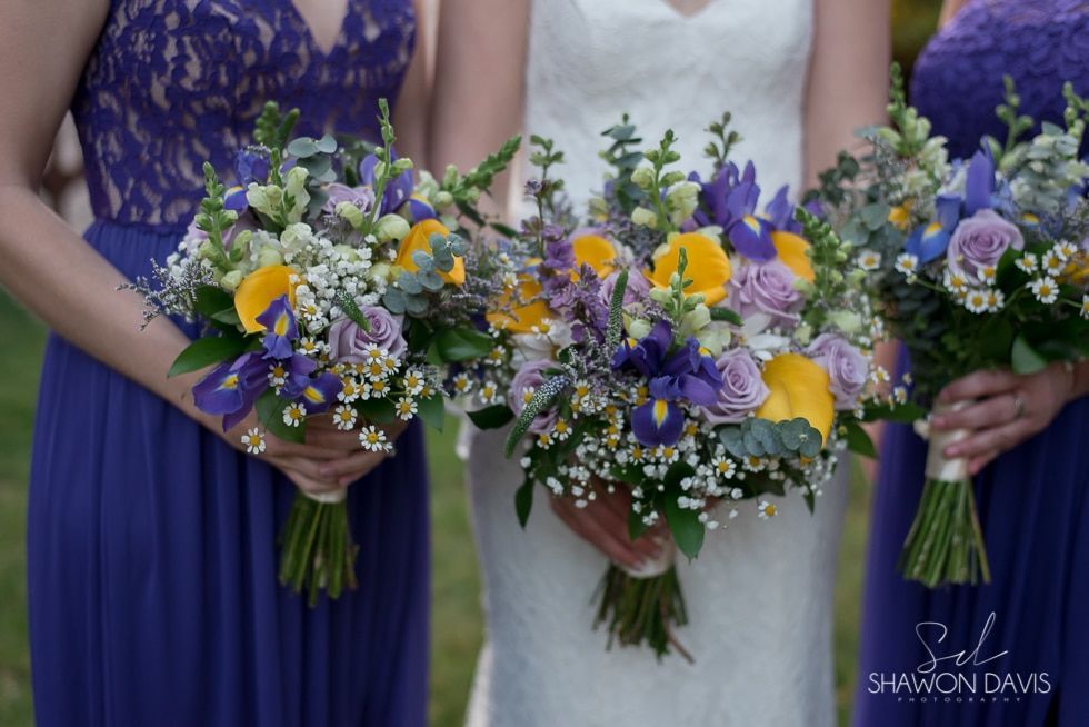 Three women hold colorful purple, yellow, and white bouquets at a wedding ceremony
