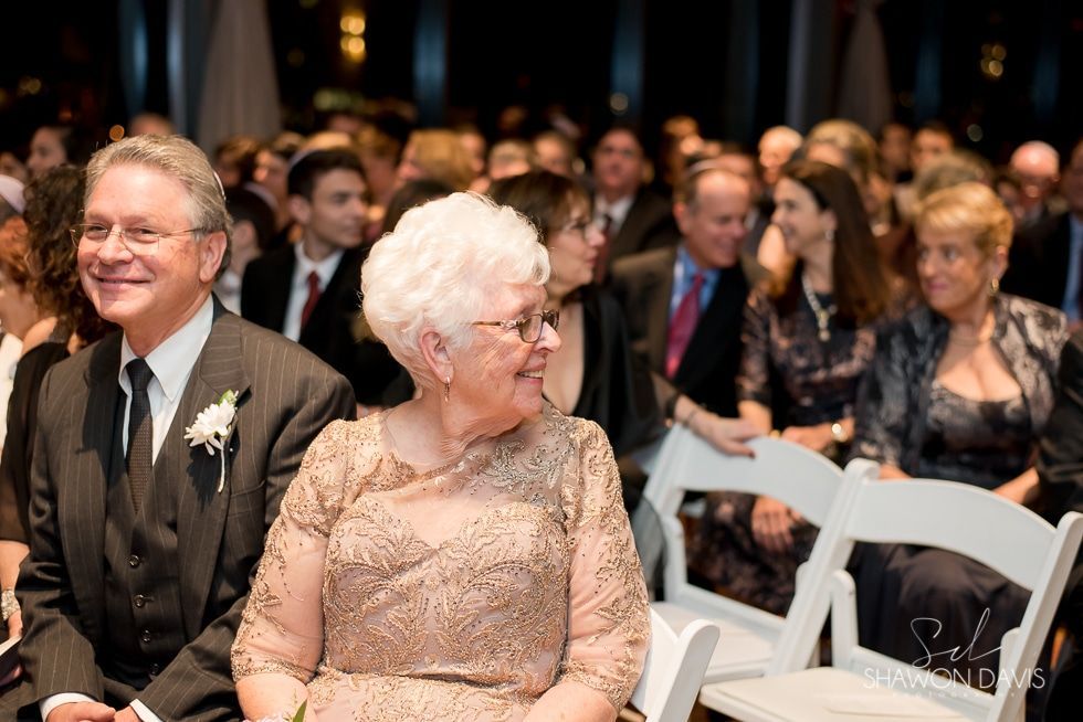 Guests seated at a formal event, smiling in front-row chairs with a crowd in the background