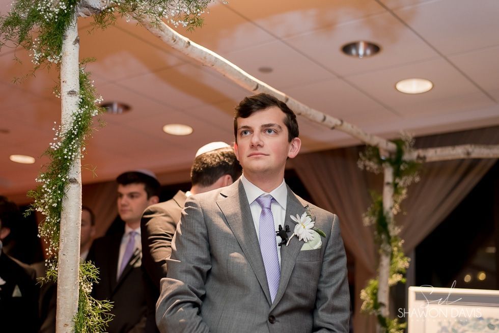 Man in gray suit with boutonniere standing at a formal indoor event under warm lighting.