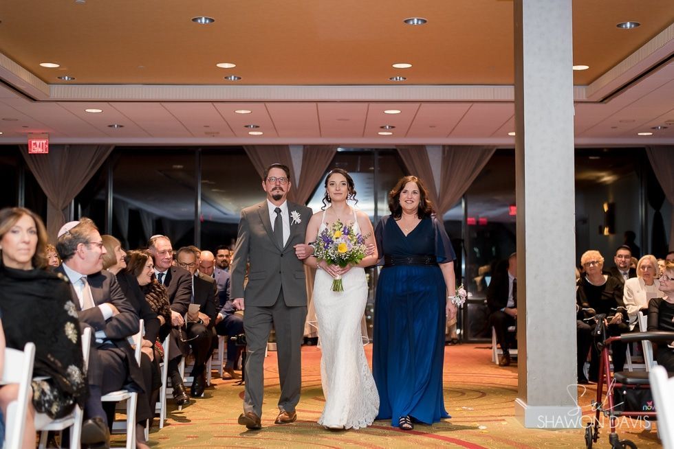 Bride enters reception hall with two companions, holding a bouquet, as guests watch from seated tables.
