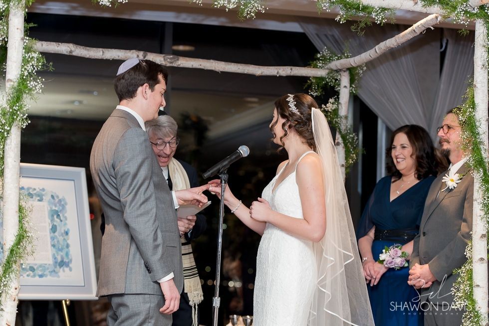 Wedding couple exchanging vows under a decorated arch, with officiant and guests watching.