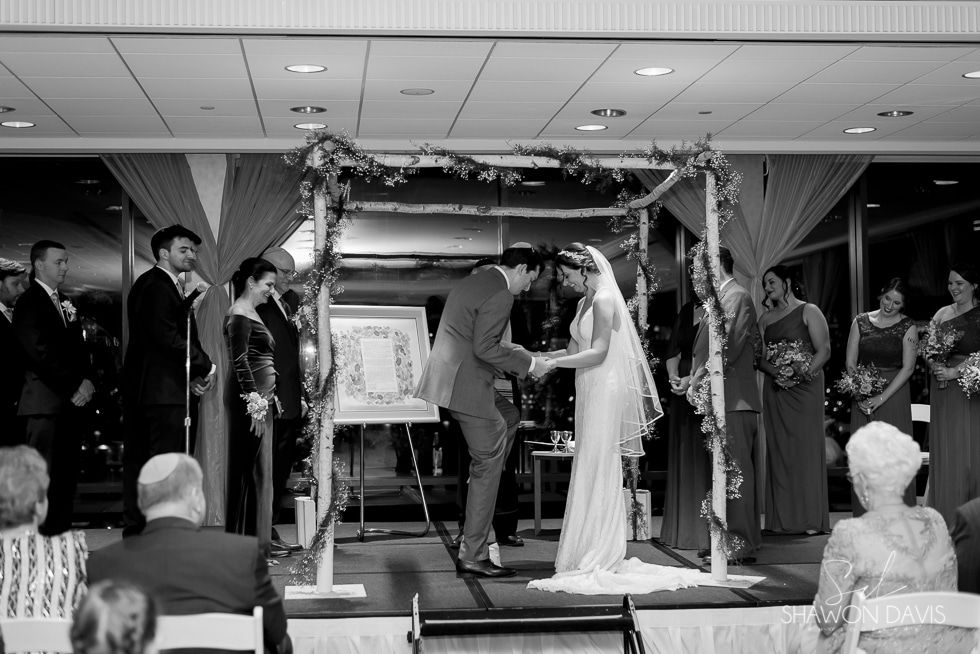 Wedding couple under decorated arch at indoor ceremony, guests seated nearby