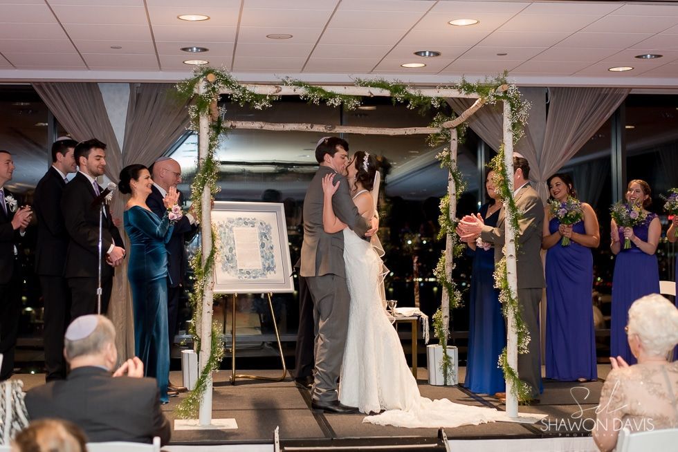 Wedding couple exchanging rings under a floral arch, surrounded by attendants at an indoor ceremony