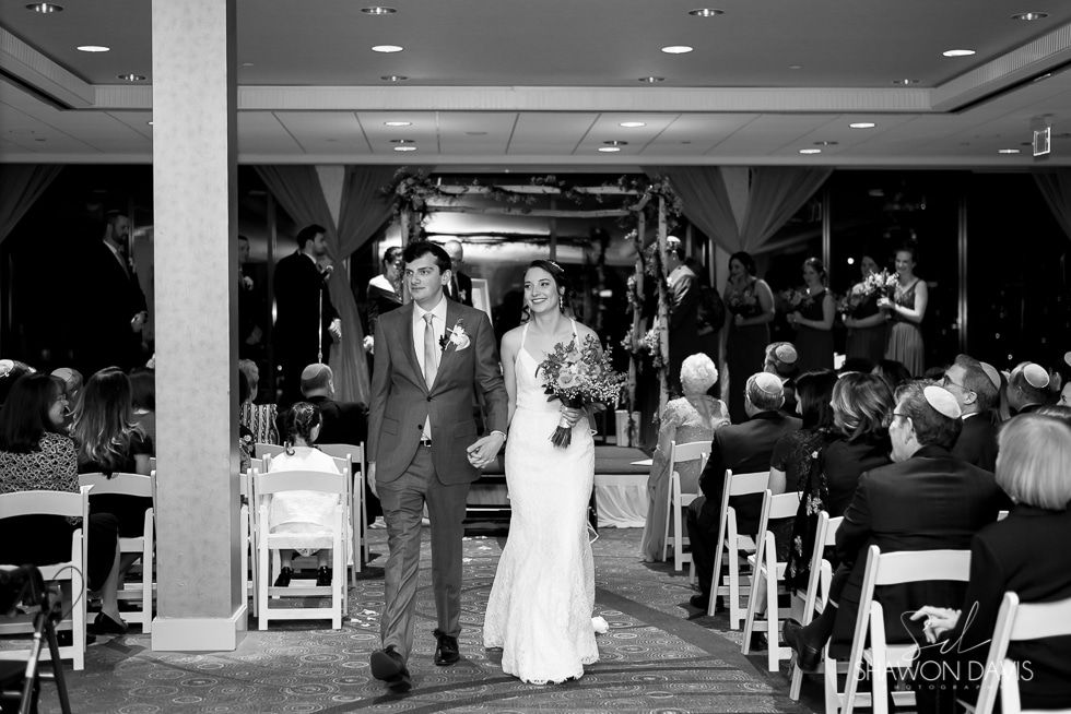 Bride and groom walking down the aisle at a wedding reception, with guests seated on both sides.