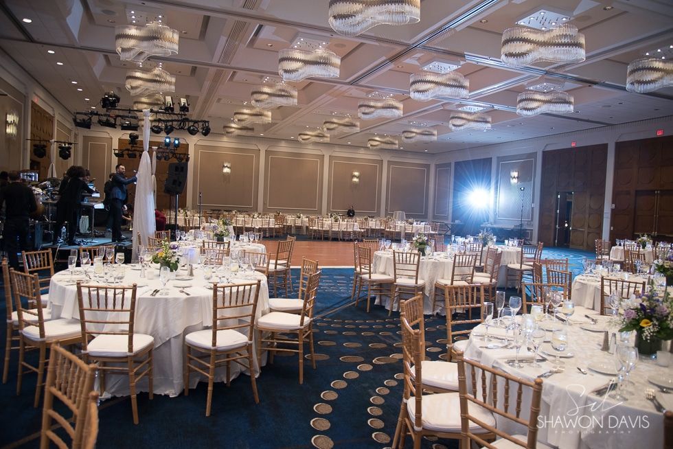 Banquet hall with round, white-clothed tables, gold chairs, and a stage with bright lights.