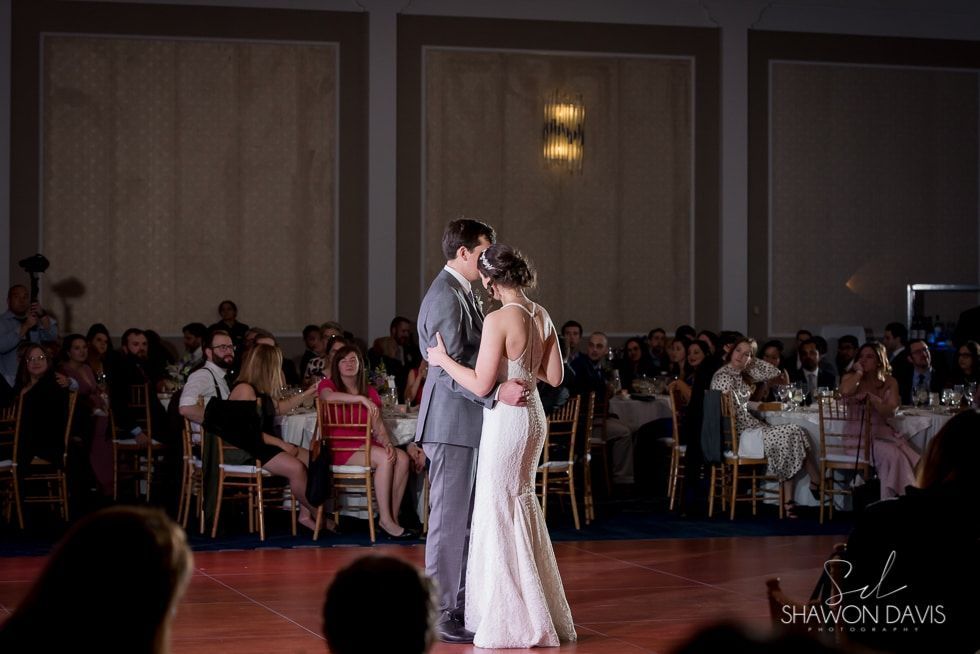 Couple slow dancing at a formal reception, surrounded by seated guests in a ballroom.