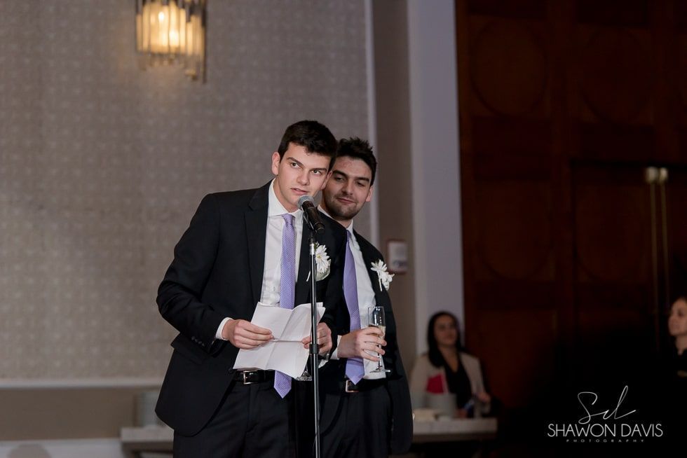 Man in a dark suit speaking at a microphone, holding papers, with another man standing behind him at a formal event.