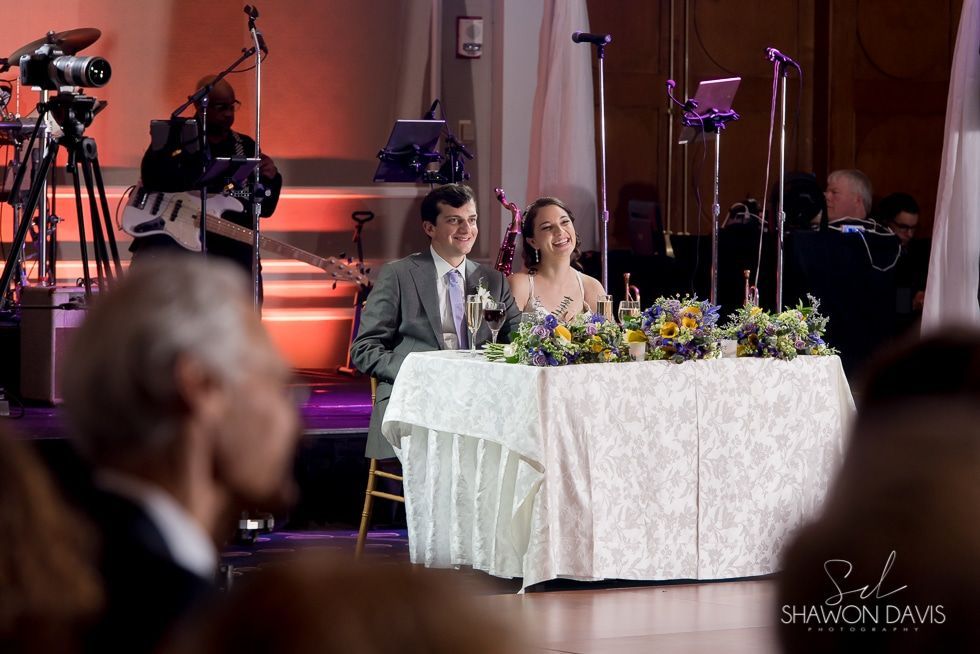 Couple seated at a decorated banquet table on a stage, with cameras and lights in the background.