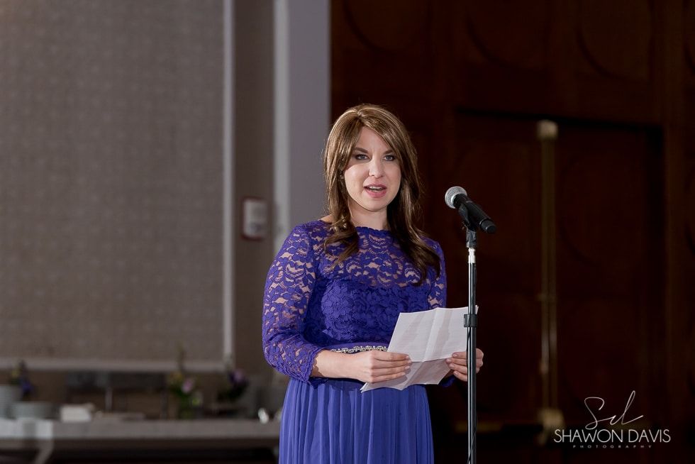 Woman in a blue dress speaking at a microphone and holding papers at an indoor event