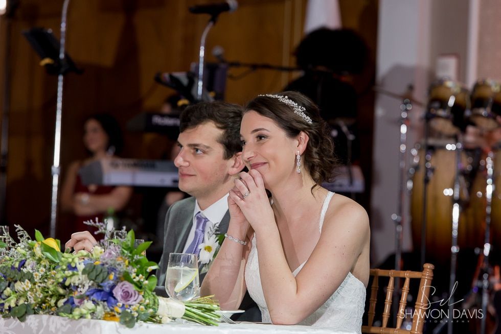 Bride and groom seated at a decorated reception table, smiling and watching the event