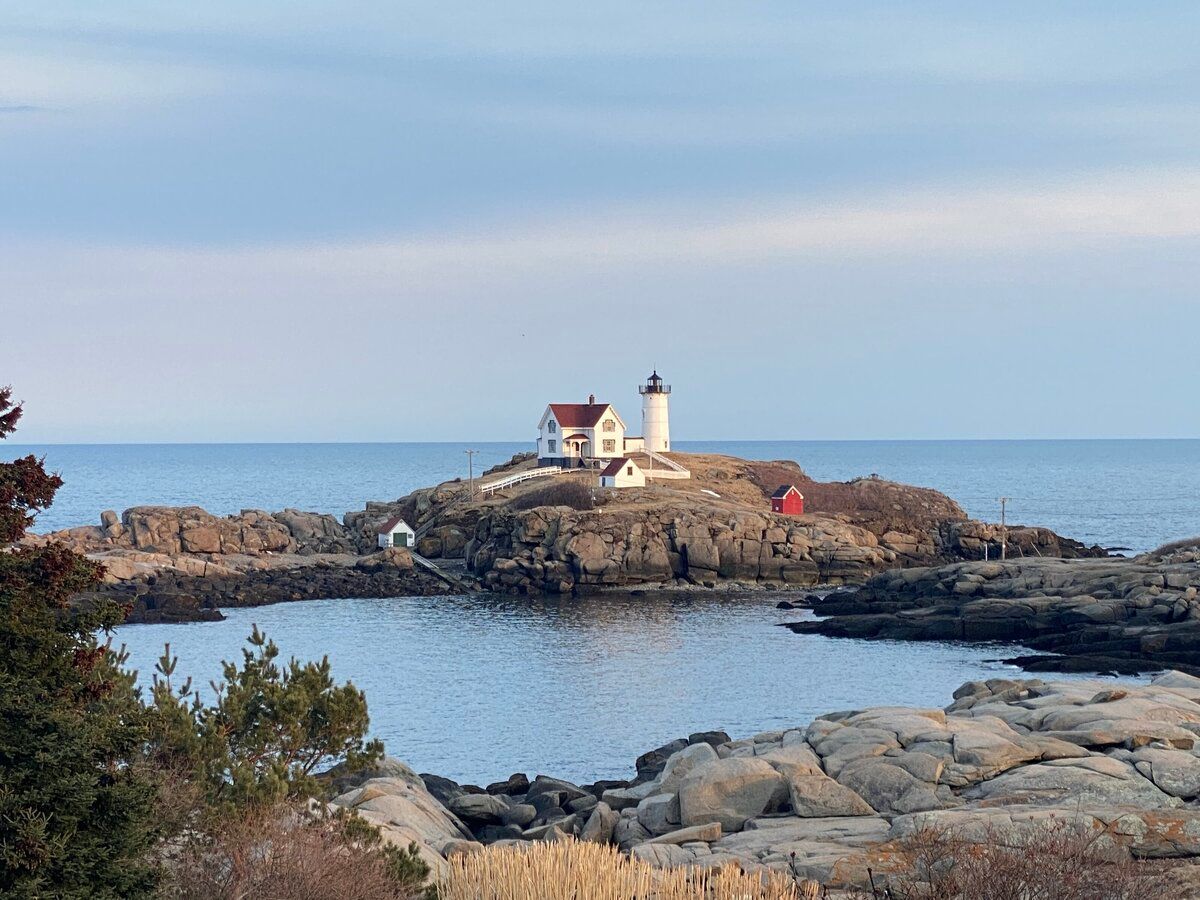 Lighthouse on rocky coastal islet at dusk, with calm water and pastel sky.