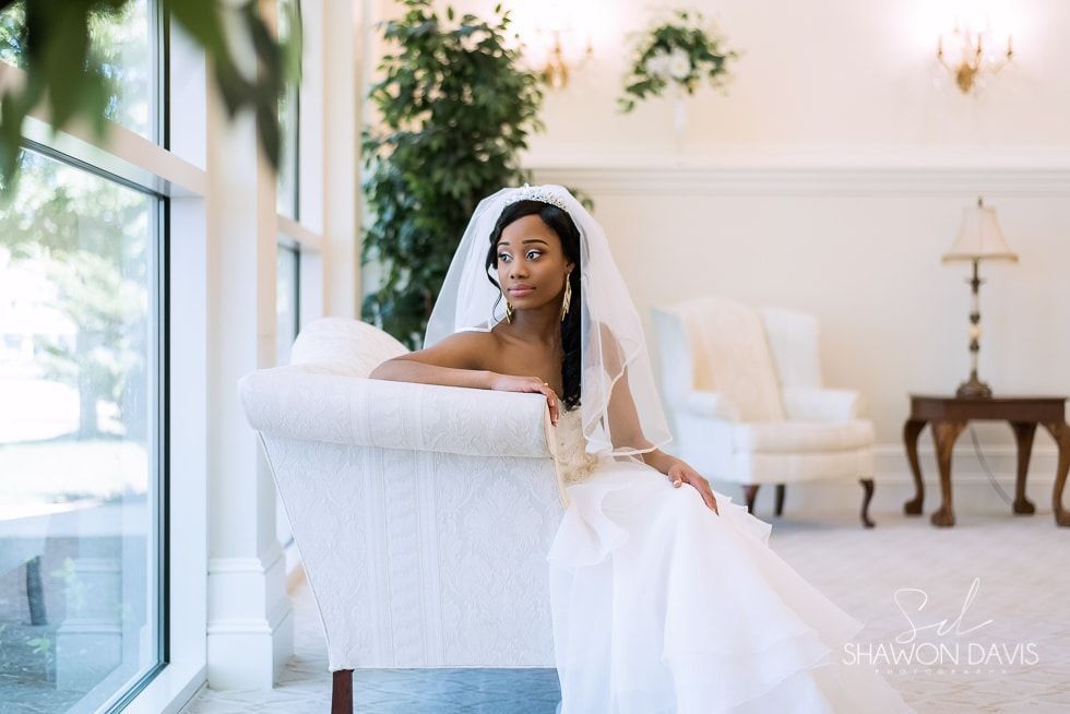 Bride in a white gown and veil sits on a white sofa in a bright room by the window.