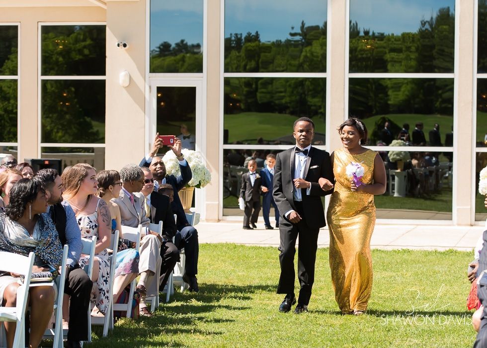 Two people walk down an outdoor wedding aisle between seated guests, in front of a glass-walled building.