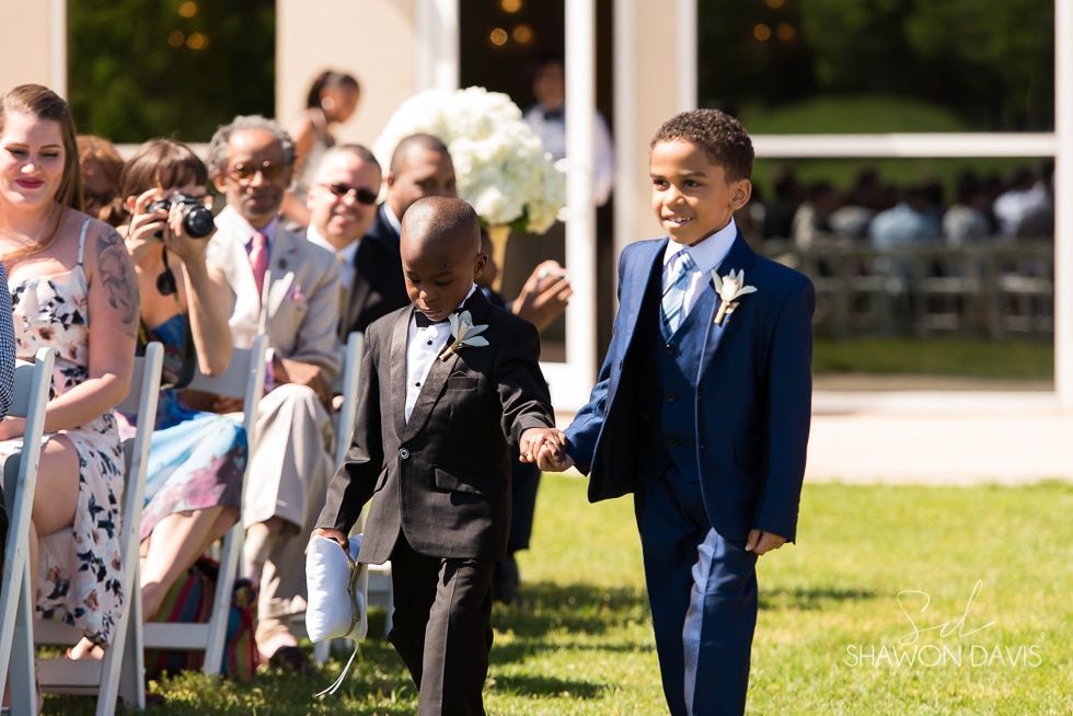 Children in suits walk hand in hand down an outdoor aisle at a sunny ceremony
