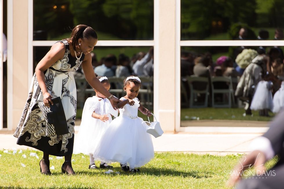 Woman helping two children in white dresses walk on grass outdoors by a building