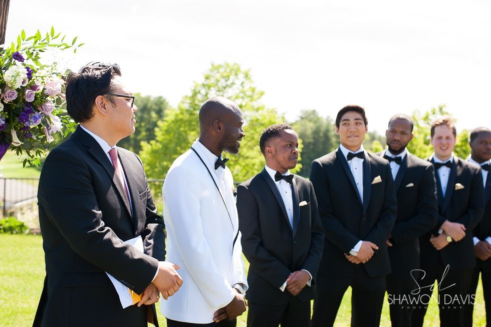 Six men in formal suits stand outdoors on a sunny lawn, facing left during a wedding procession.