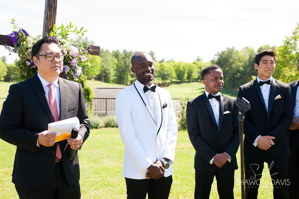 Four men in suits standing outdoors at a formal event, one holding papers and flowers.
