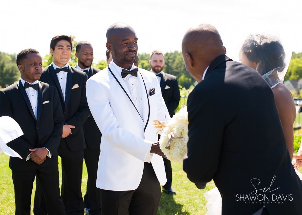 Wedding party in tuxedos outdoors, with a man in a white jacket greeting another man in a black suit.