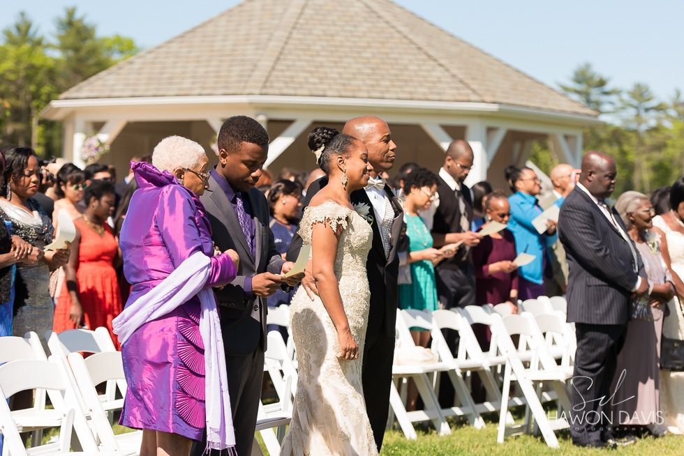 Guests walking outdoors at a wedding ceremony, with white chairs and a gazebo in the background