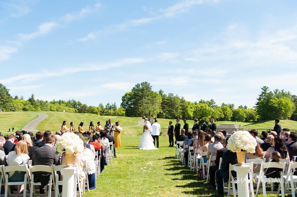 Outdoor wedding ceremony with guests seated on white chairs along a grassy aisle under a blue sky