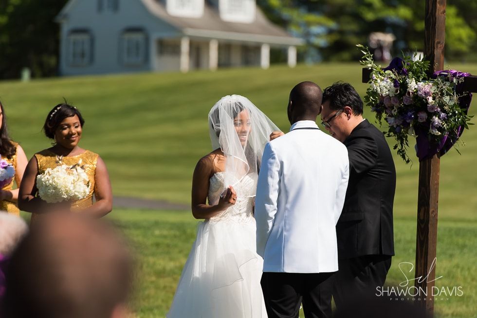 Bride and groom exchanging vows outdoors, with a bridesmaid nearby and a floral arch at the ceremony.