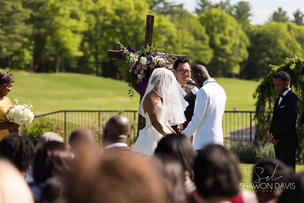 Bride and groom kiss at outdoor wedding ceremony with guests and floral arch in a sunny park